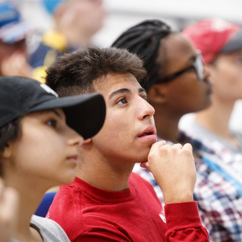 A student looking up with his hand on his chin sitting with a crowd of students.