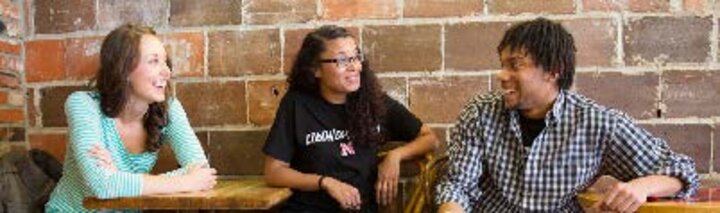 Students talking with each other while seated at tables in front of a brick wall.